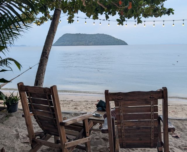 Zwei Holzstühle am Strand unter Bäumen mit Lichterkette, Blick aufs Meer und eine kleine Insel
